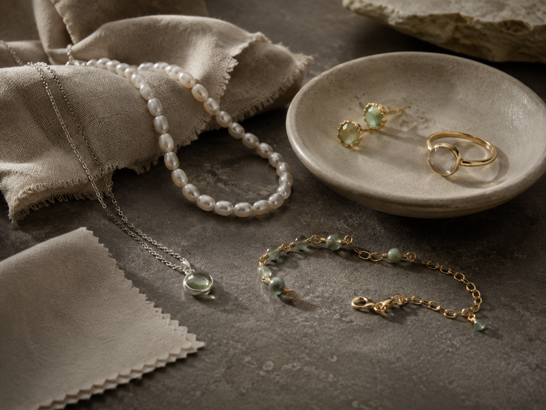 A pearl necklace, natural stone earrings, rings, and bracelets are displayed on a warm gray stone surface, accompanied by linen cloth and jewelry polishing cloths, creating an understated, natural atmosphere for jewelry care.