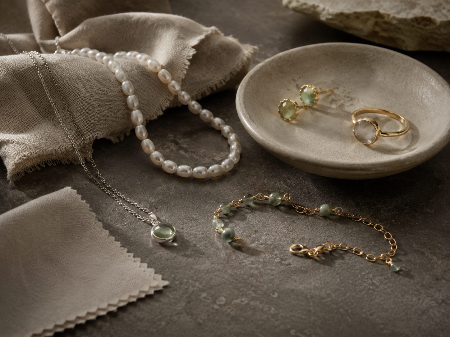 A pearl necklace, natural stone earrings, rings, and bracelets are displayed on a warm gray stone surface, accompanied by linen cloth and jewelry polishing cloths, creating an understated, natural atmosphere for jewelry care.
