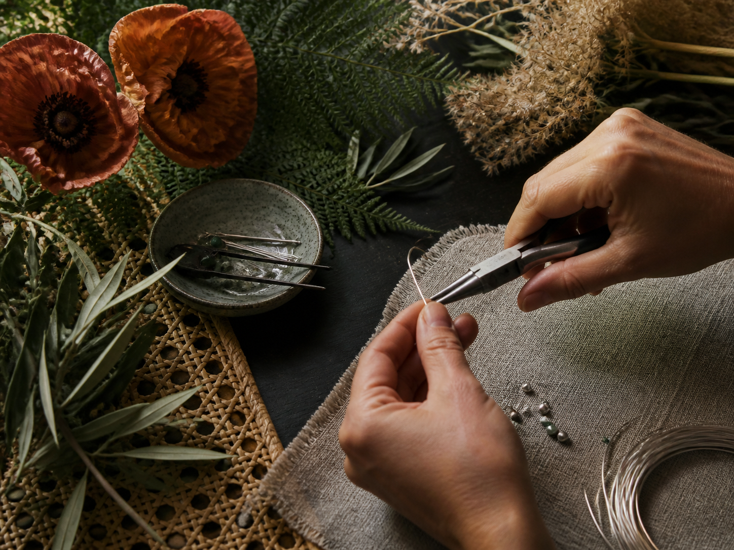 Hands shaping silver wire among poppies, fern and woven natural materials